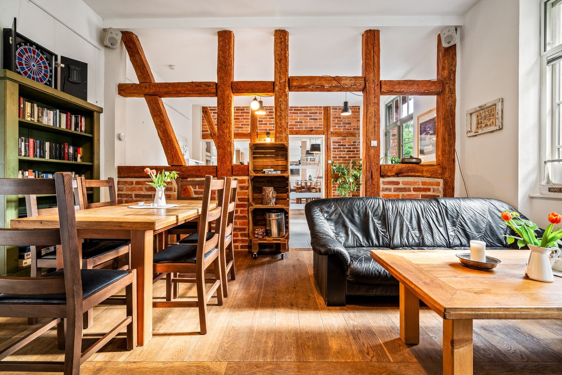 Gemütliches Loft-Wohn- und Esszimmer mit Holzbalken, schwarzem Sofa und Holztischen in der Nähe heller Fenster
