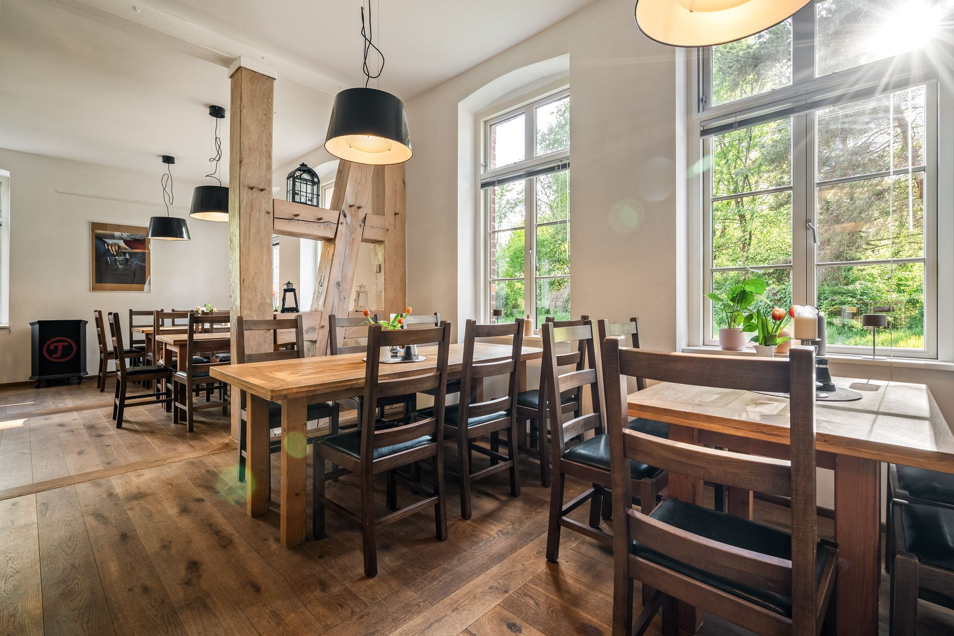 Heller Café-Essbereich mit Holztischen, Stühlen, Pendelleuchten und großen Fenstern mit Blick ins Grüne