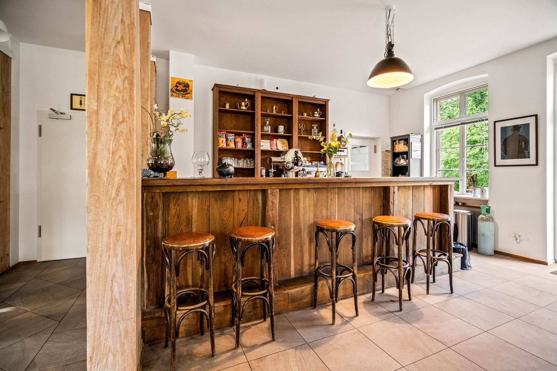 Rustikale Küche mit Holzbar, Barhockern, Regalen und Pendelleuchte, neben hellen Fenstern und Fliesenboden