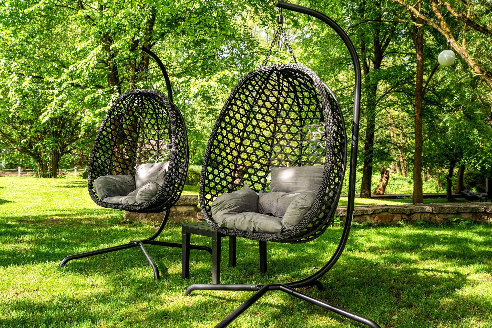 Zwei hängende Korbsessel in Eierform mit grauen Kissen in einem sonnigen grünen Park