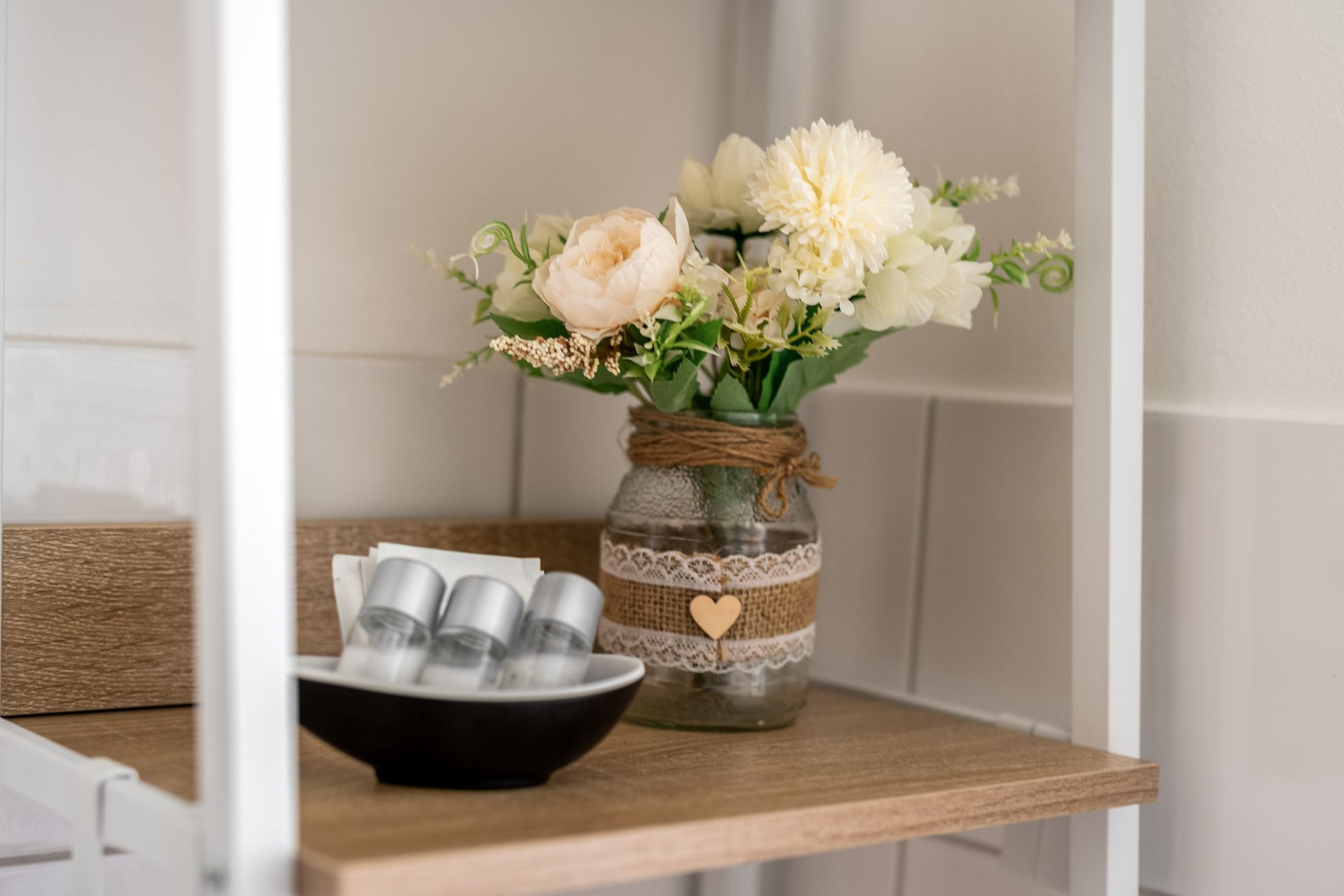 Ein rustikaler Tisch mit einer Vase weißer Blumen und einem schwarzen Tablett mit silbernen Bechern auf einem hellen Eckregal.