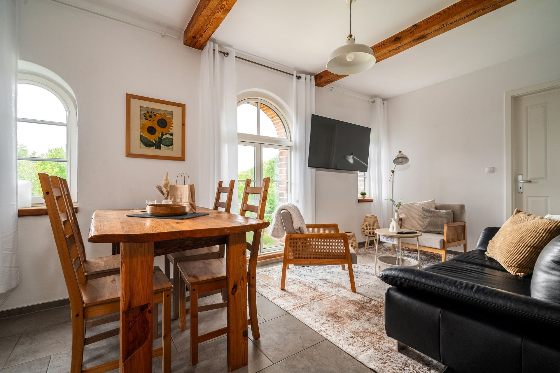 Helles Wohn-Esszimmer mit Holzbalken, Tisch, Korbstühlen, Sofa, Fernseher und Bogenfenstern.
