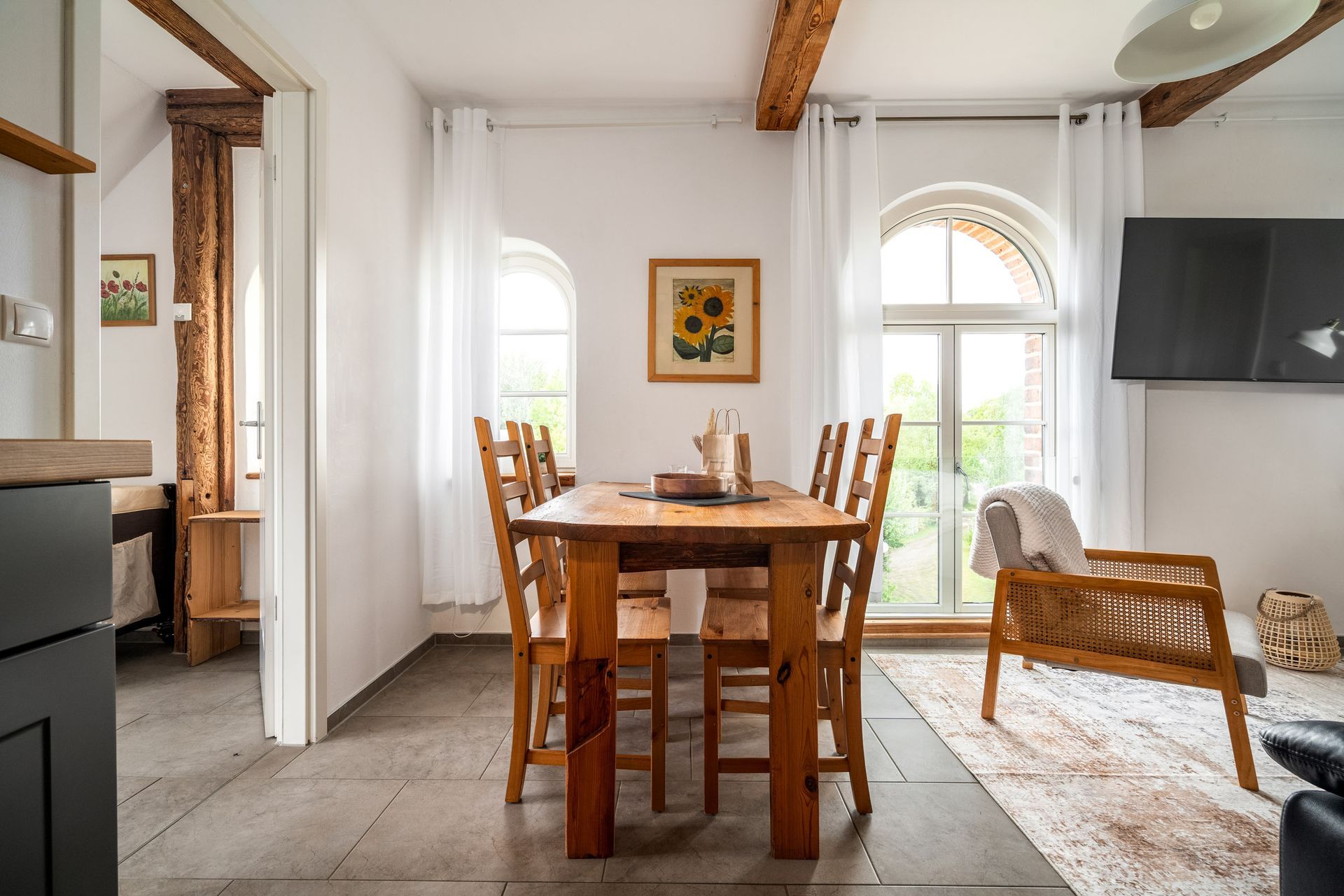 Helles Esszimmer mit Holztisch, Stühlen, großen Fenstern und an der Wand montiertem Fernseher