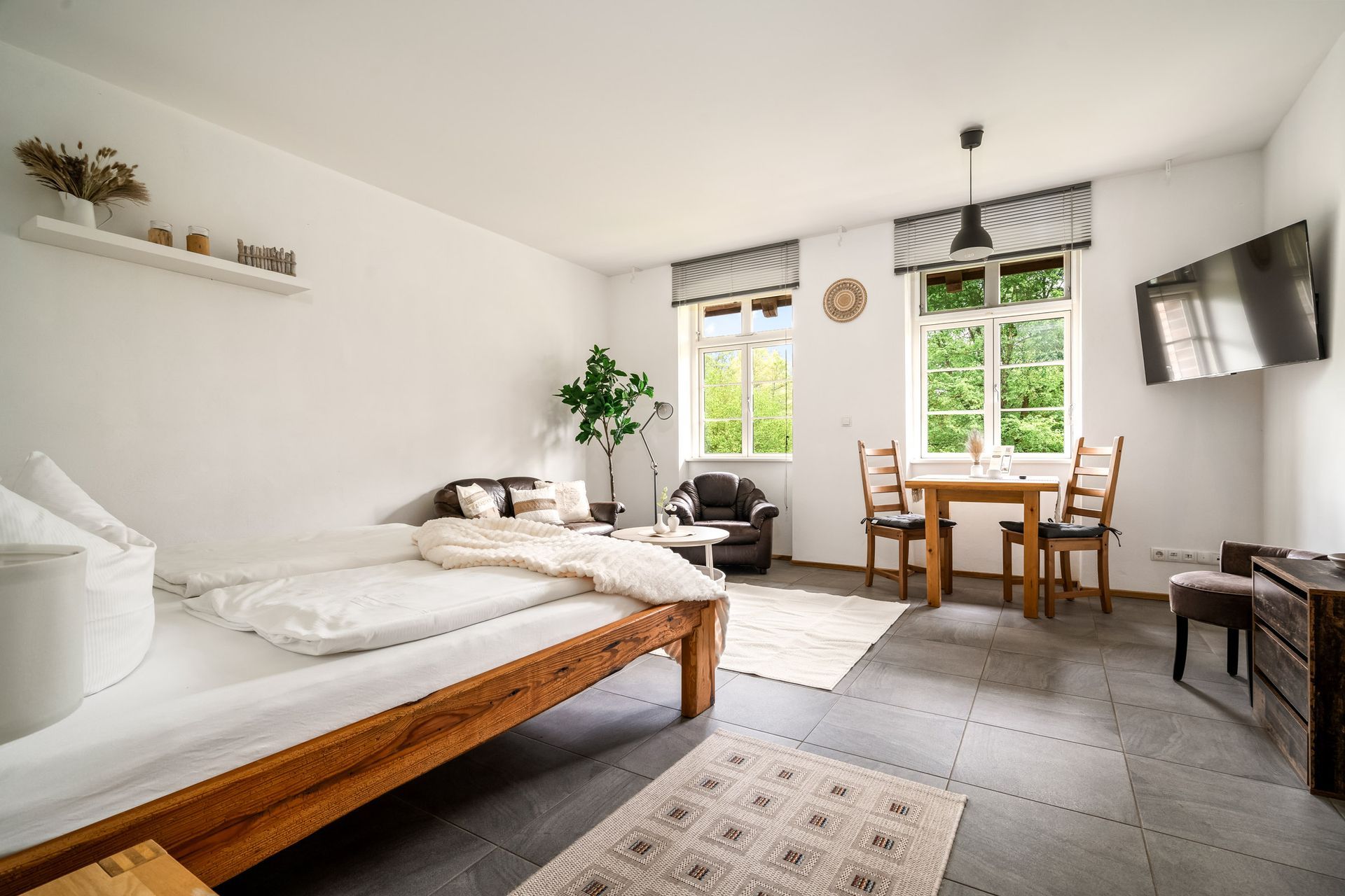 Helles, minimalistisches Schlafzimmer mit weißen Wänden, Holzbett, Schreibtisch am Fenster und an der Wand montiertem Fernseher