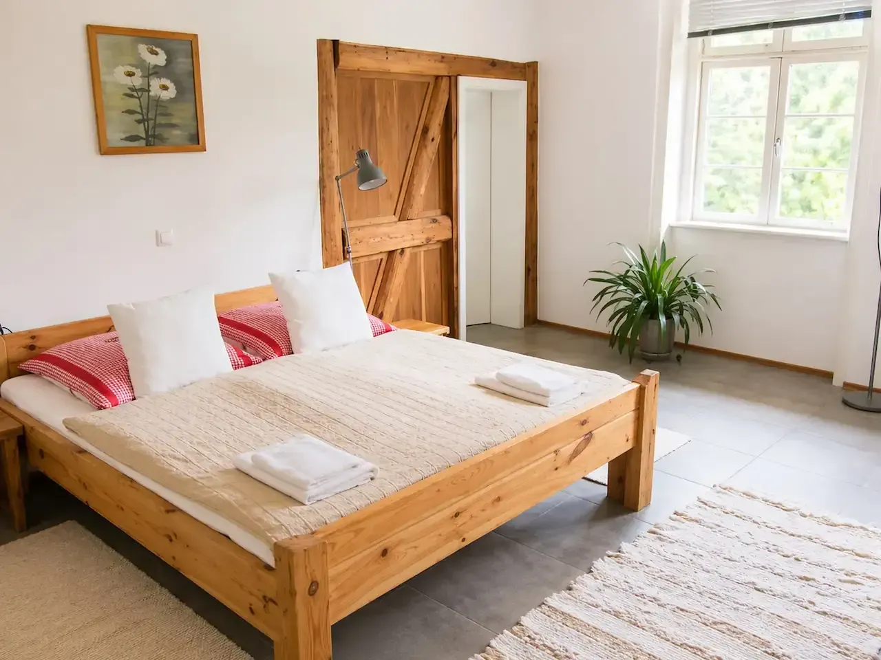 Helles Schlafzimmer mit Holzbett, weißer Bettwäsche, roten Kissen, Fenster, Pflanze und gewebtem Teppich