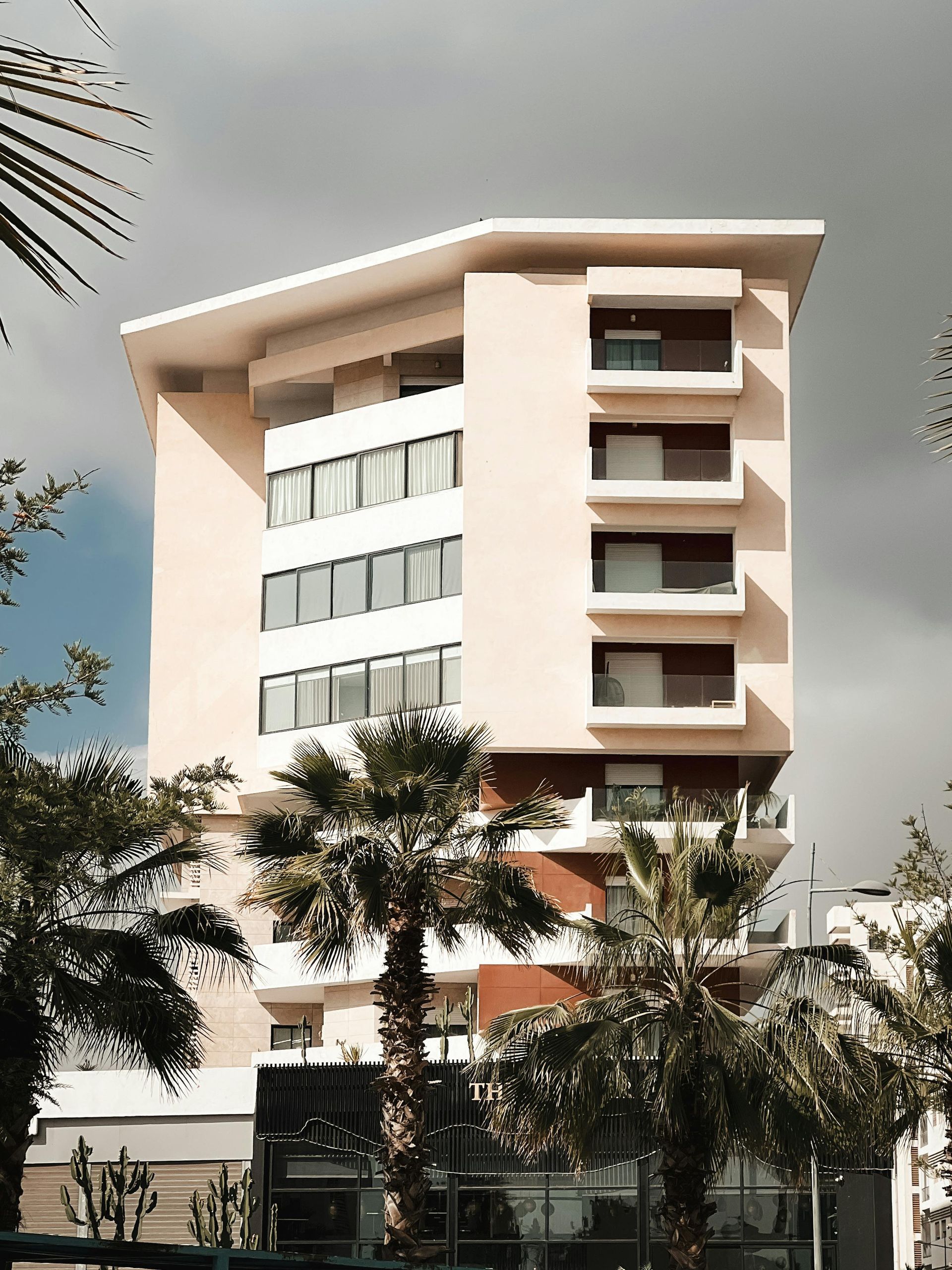 Edificio de apartamentos moderno de color beige y blanco con árboles al frente bajo un cielo nublado.