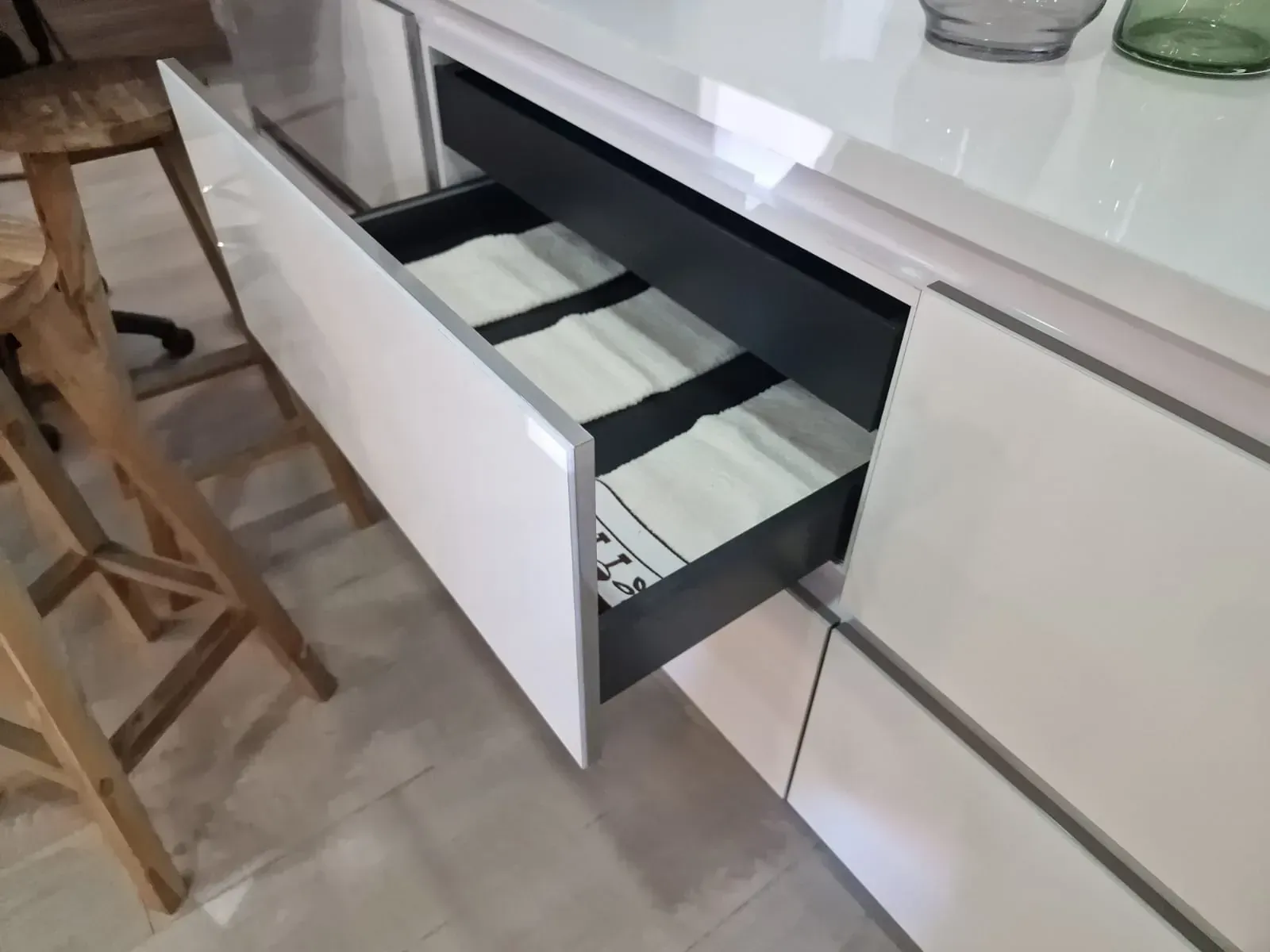 Cajón de cocina abierto con utensilios en su interior, junto a un taburete y una encimera.