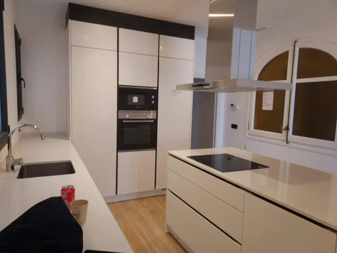 Cocina moderna blanca con isla, horno empotrado y grandes ventanales.