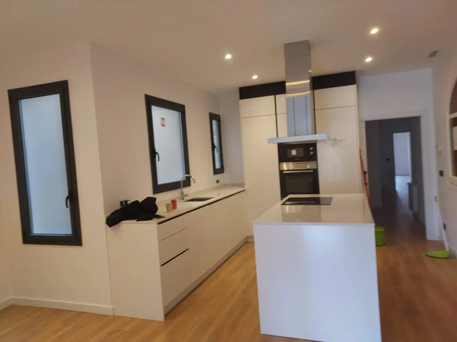 Cocina moderna blanca con isla, suelos de madera, horno empotrado y luces empotradas en el techo.