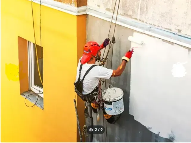Un trabajador pinta el exterior de un edificio desde una plataforma suspendida, aplicando pintura gris junto a una pared de color amarillo brillante.