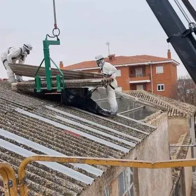 Trabajadores levantando tejas con una grúa en un tejado inclinado junto a un edificio de ladrillos.