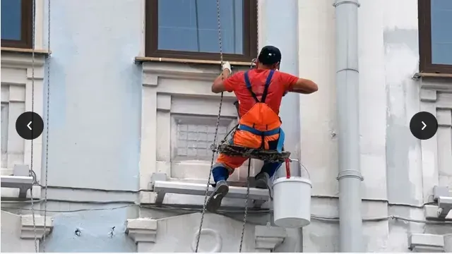 Un trabajador con arnés de seguridad naranja pinta la fachada de un edificio en un andamio cerca de una ventana.