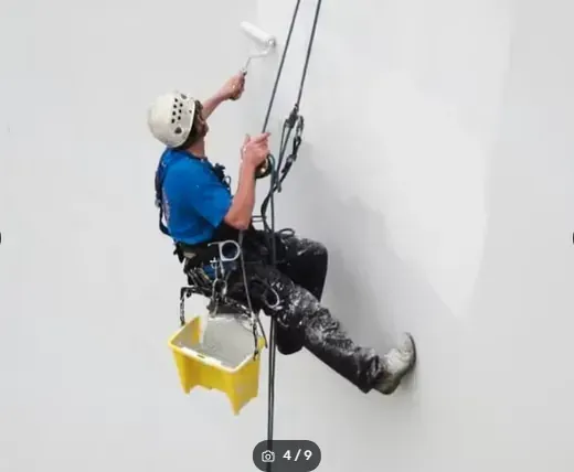 Escalador con casco y arnés descendiendo en rápel por una pared blanca con cuerdas y un cubo amarillo debajo.
