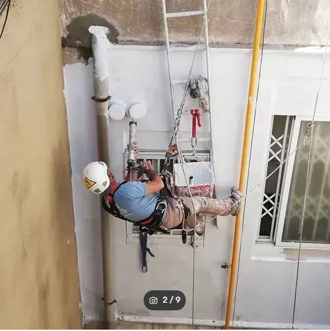 Un trabajador, suspendido de una cuerda en el exterior de un edificio, repara las tuberías cerca de una ventana.