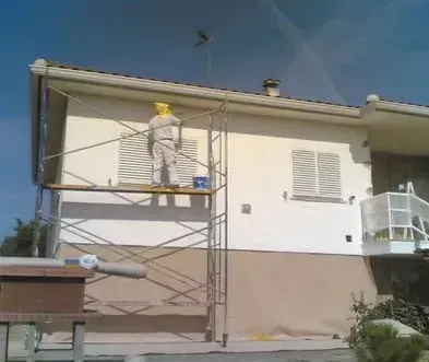 Un trabajador con mono blanco y casco amarillo pinta el exterior de una casa desde un andamio.