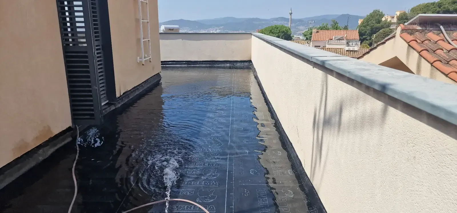 Estrecha terraza en la azotea con baldosas negras en el suelo, paredes blancas y vistas a tejados y colinas en la distancia.