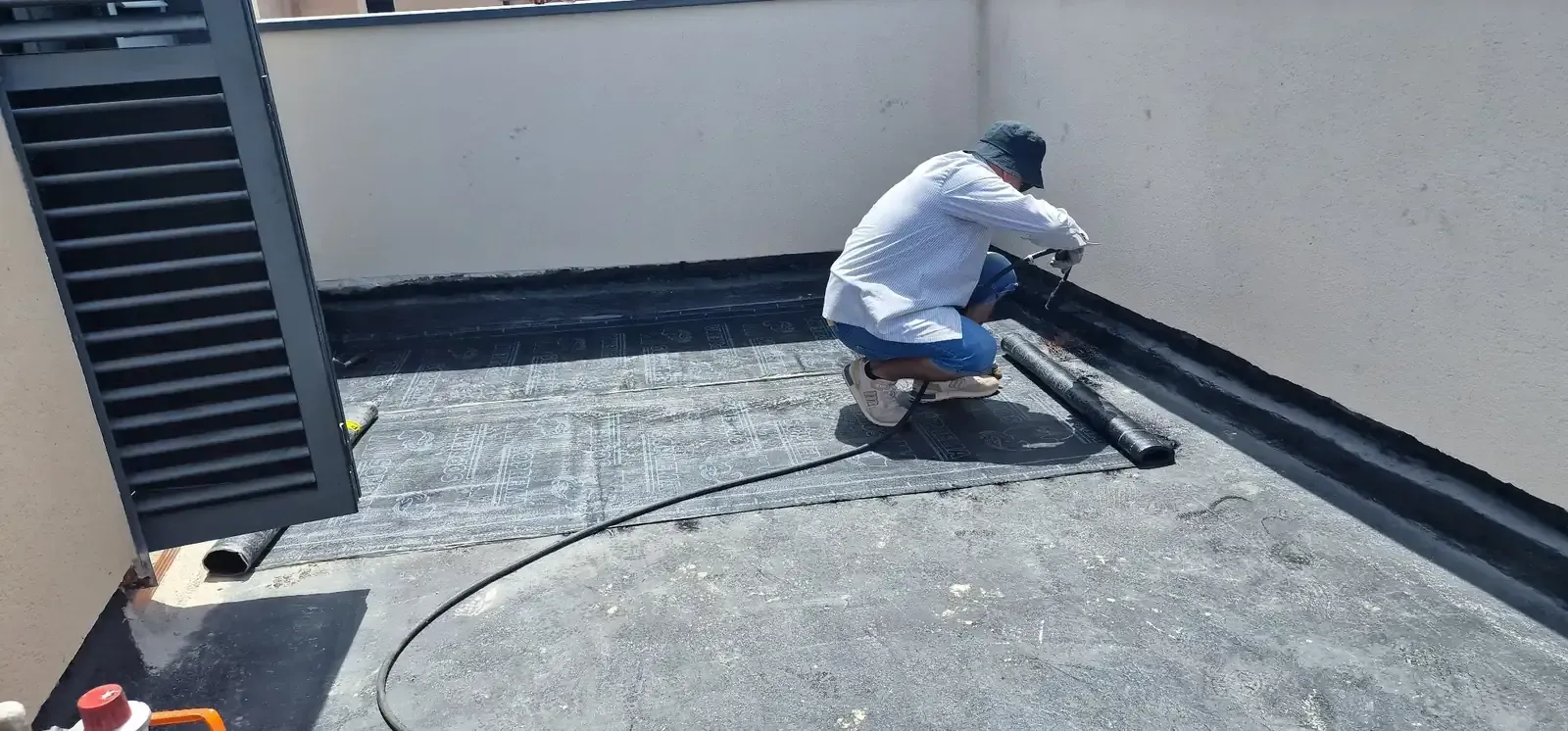 Un trabajador arrodillado en un tejado aplica impermeabilizante negro a lo largo del borde, cerca de una pared.