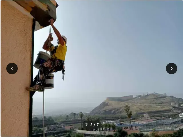 Un escalador con camisa amarilla asciende por la pared de un edificio sobre un paisaje urbano en la ladera de una colina.
