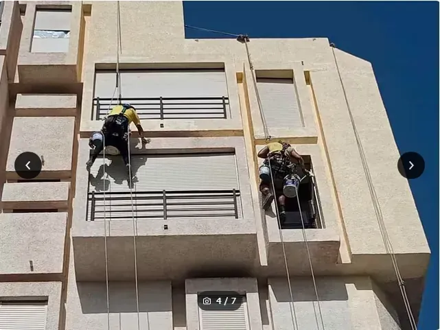 Unos trabajadores, equipados con trajes de seguridad, descienden en rápel por la fachada de un edificio de color beige utilizando cuerdas y escaleras.