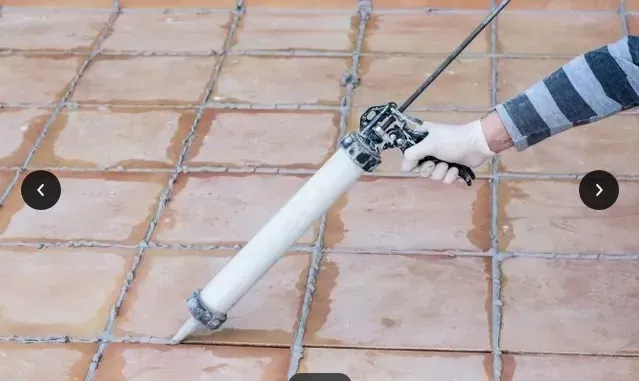 Mano sosteniendo una pistola de calafateo blanca aplicando sellador a baldosas de terracota.