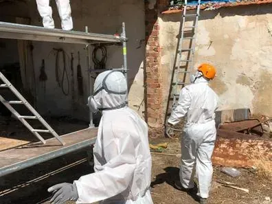 Trabajadores con trajes protectores blancos inspeccionan el exterior dañado de un edificio utilizando escaleras y escombros.