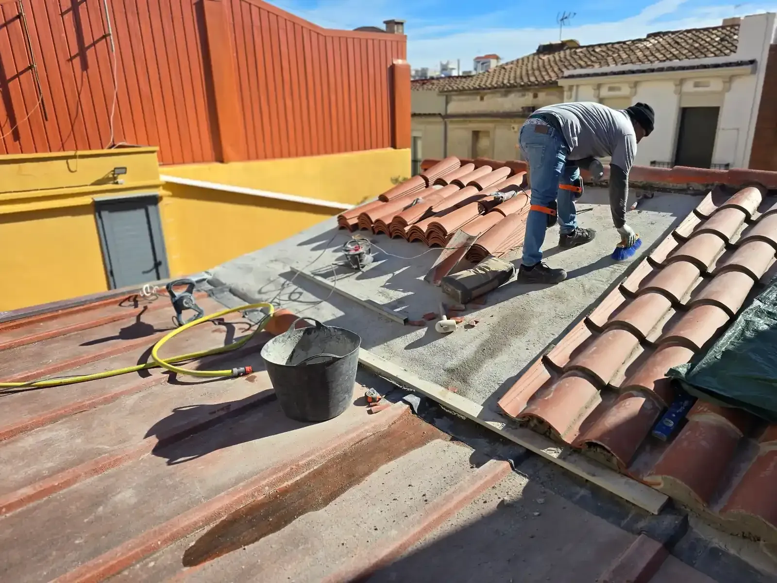 Trabajador reparando tejas de terracota en un tejado inclinado con herramientas y cubos.