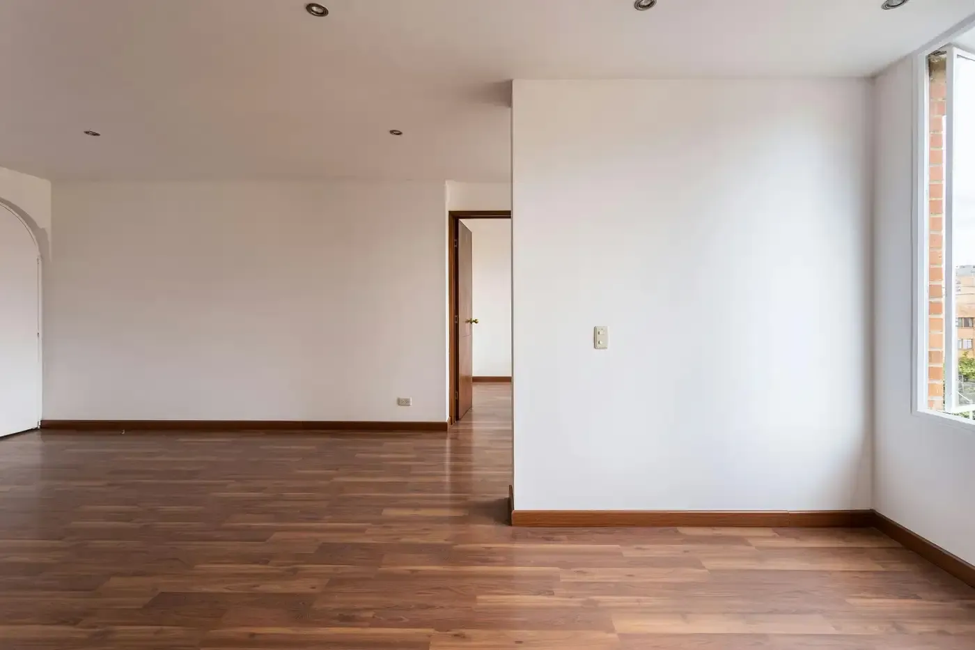Sala de estar vacía con pisos de madera oscura, paredes blancas, puerta y ventana grande.