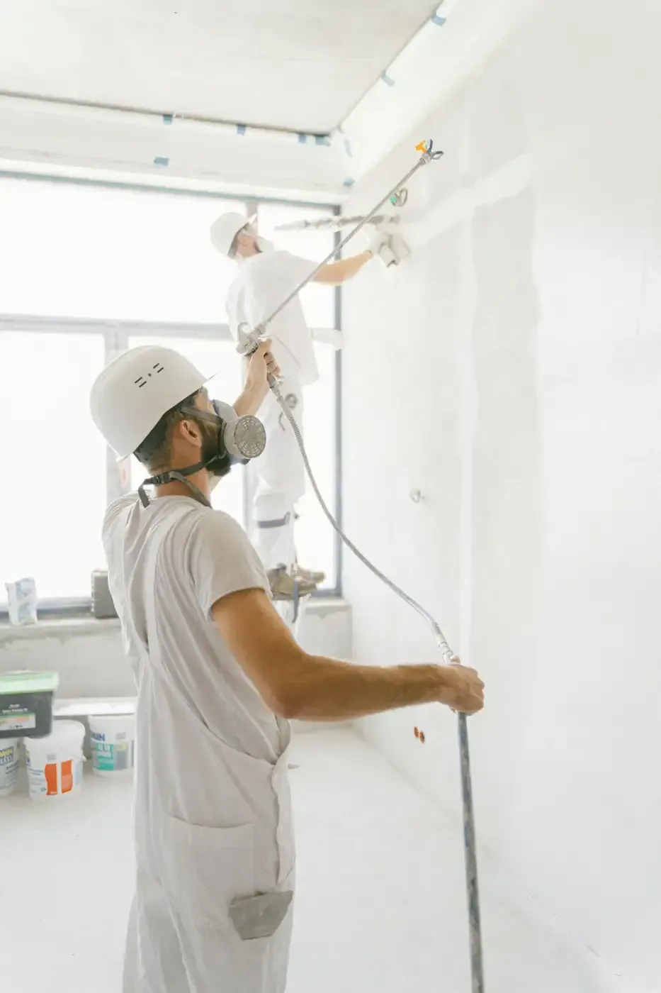 Dos trabajadores con cascos blancos pintando una pared interior de color blanco brillante sobre un andamio.
