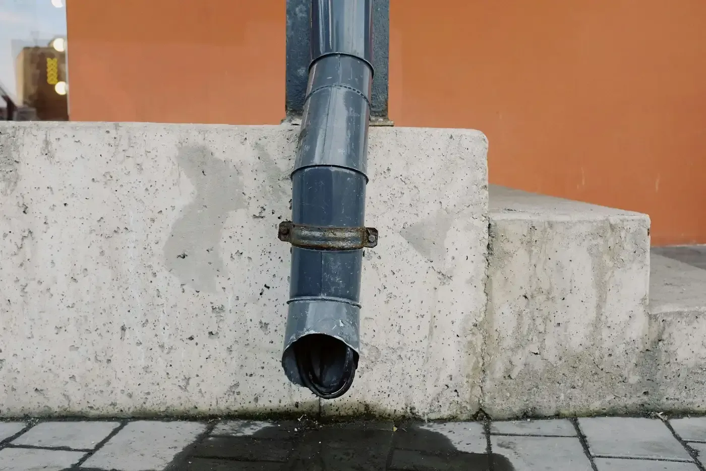 Bajante gris que termina sobre el pavimento, sujeta a una pared de hormigón con fondo naranja.