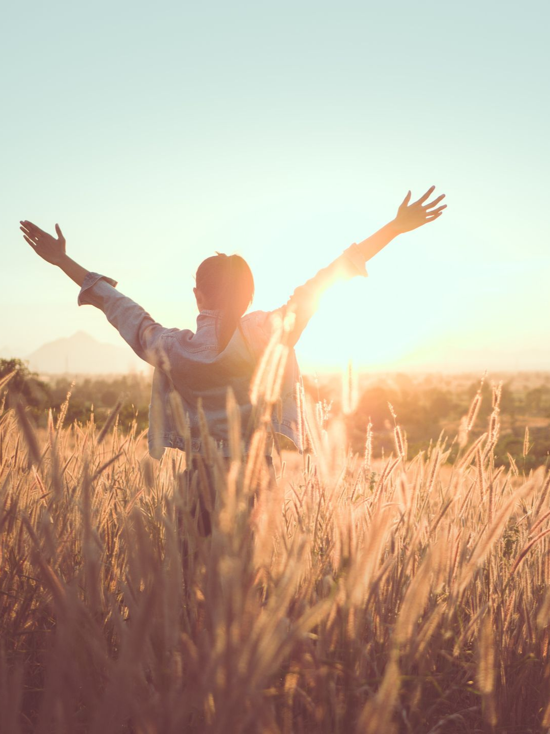 Personne aux bras tendus dans un champ d'herbes hautes, face au soleil, baignée de lumière dorée.