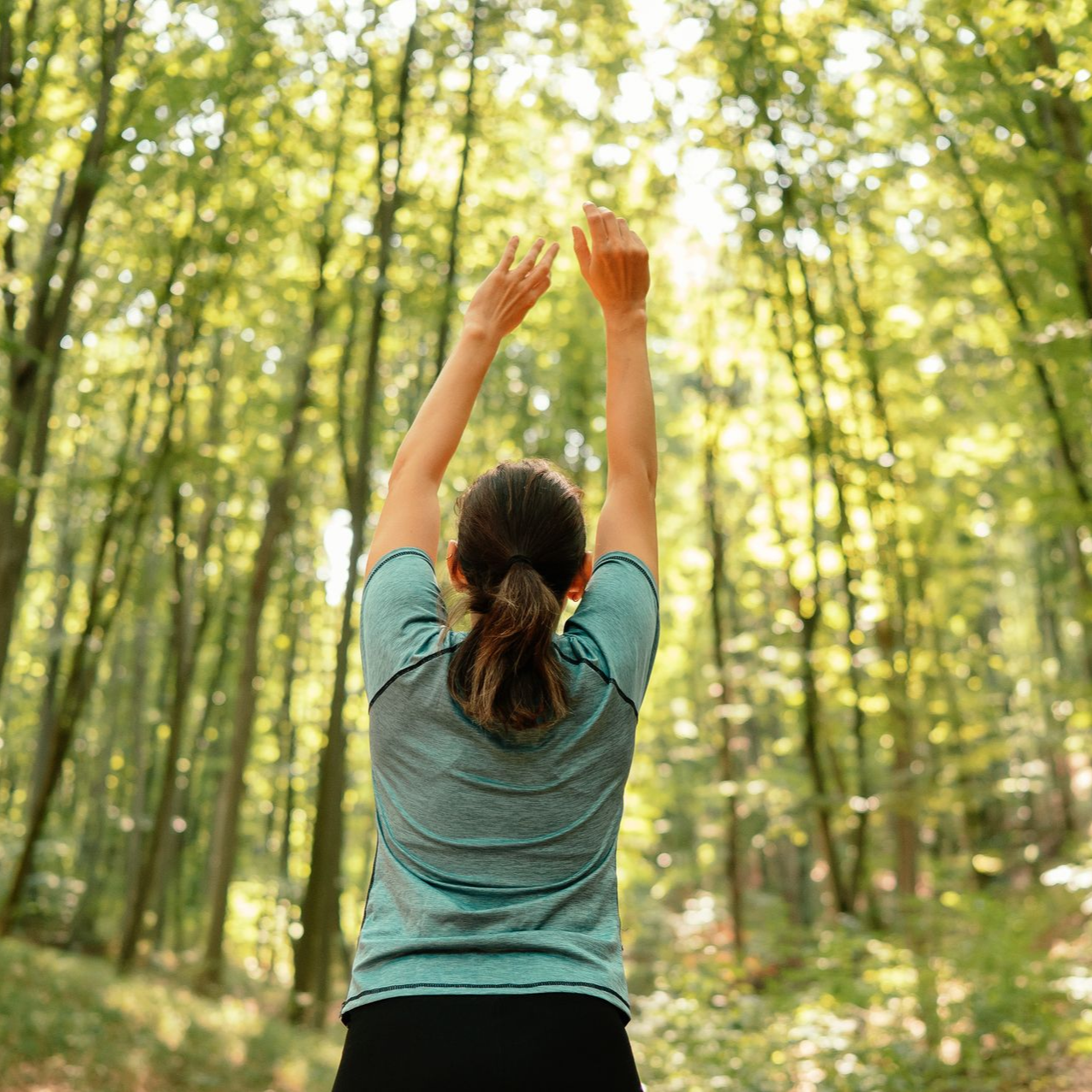 Femme aux bras levés dans une forêt, baignée de soleil.