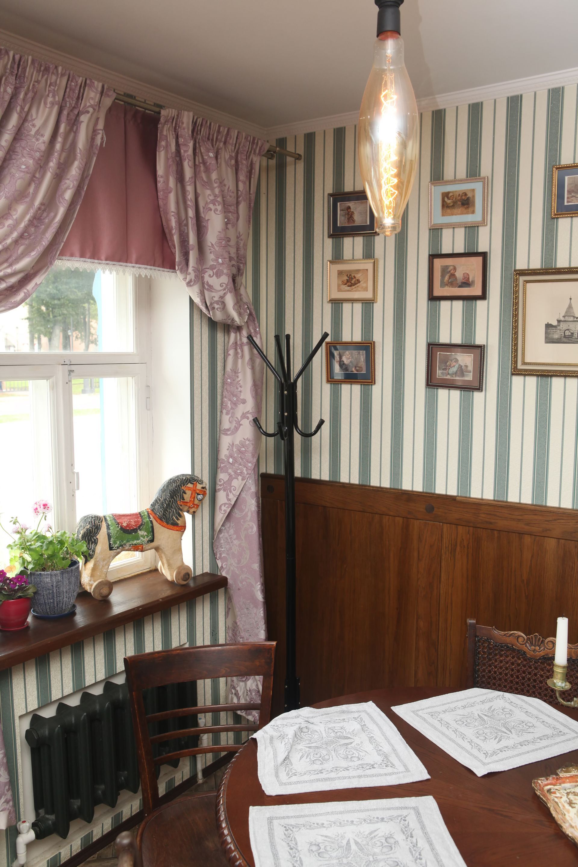 Chambre cosy avec papier peint à rayures, table ronde, décoration antique, fenêtre avec rideaux et lampe décorative.