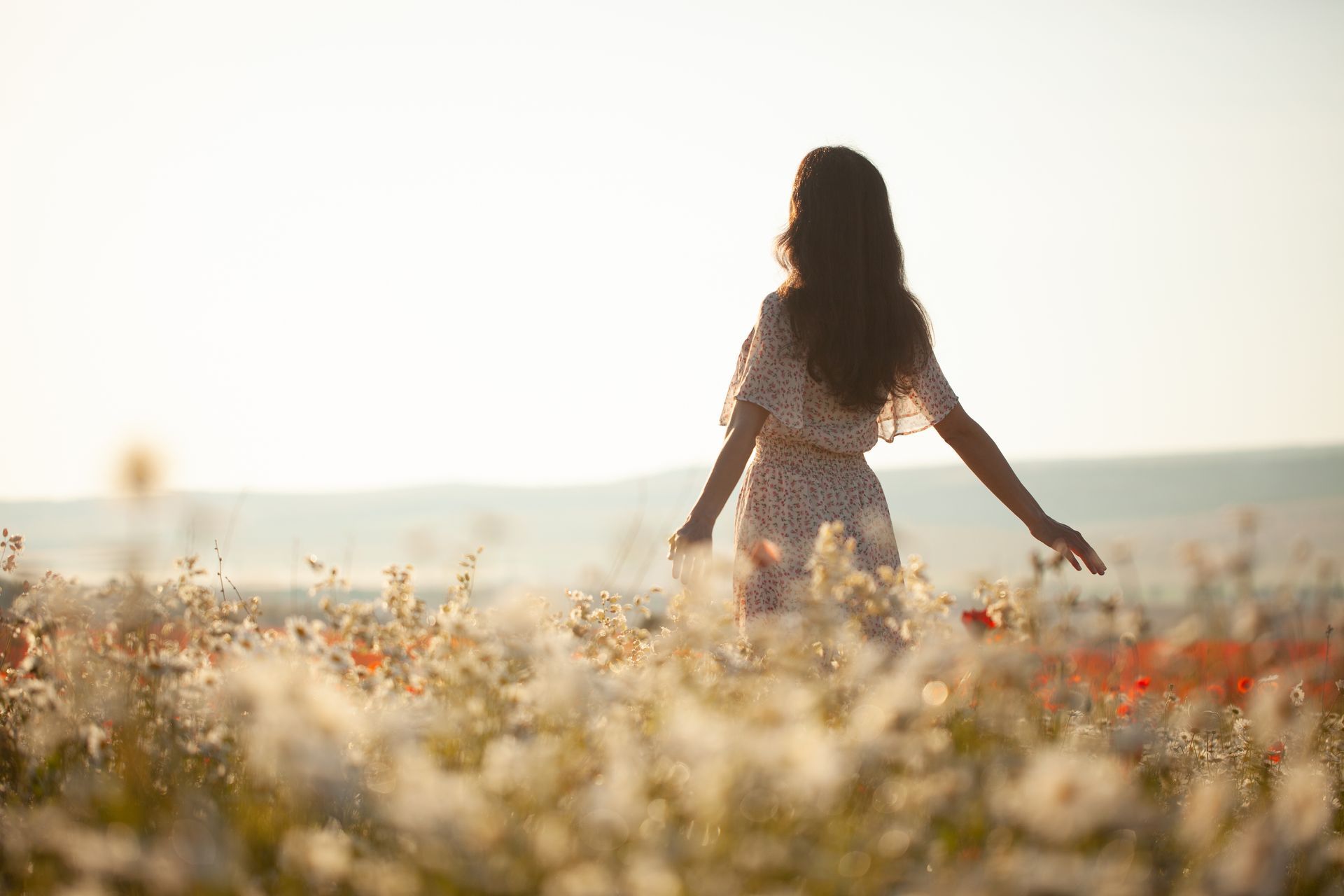 Femme en robe fleurie marchant dans un champ de fleurs ensoleillé, les bras tendus.