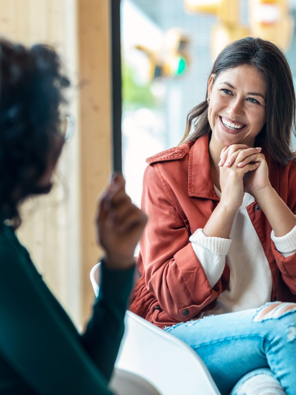 Femme avec un grand sourire et les mains jointes, assise en face d'une personne dans un café ; fenêtre et rue visibles.
