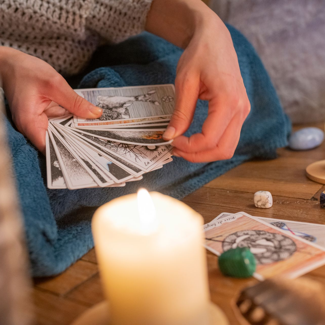 Personne mélangeant des cartes de tarot avec une bougie allumée, des cristaux et des dés sur une surface en bois.