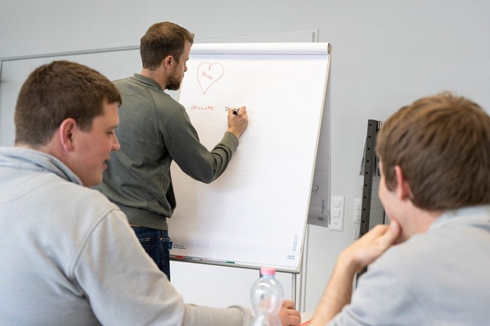 Person mit Stift in der Hand steht vor Gruppe, die auf Stühlen sitzt
