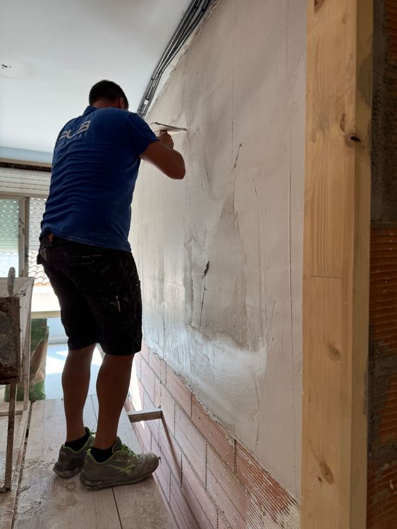 Tres pintores en escaleras trabajan en una pared interior, uno usando un pincel y dos aplicando pintura.