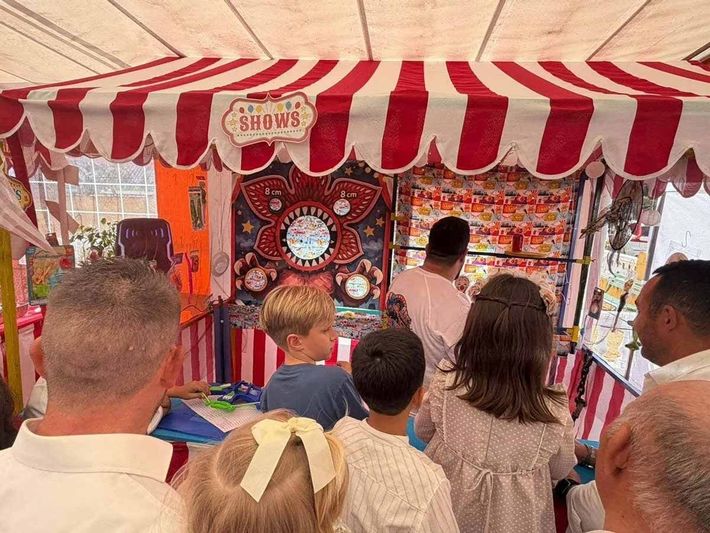 Gente observando un juego de feria bajo una carpa de rayas rojas y blancas, con una colorida pared de premios y un tablero de tiro al blanco.