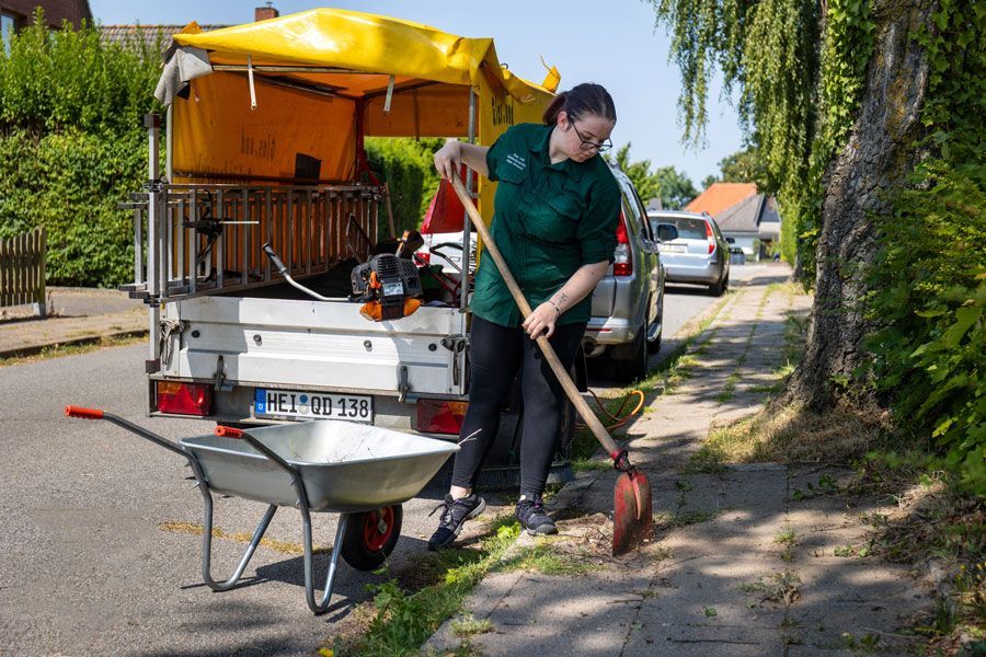 Eine Person harkt Laub neben einer Schubkarre und einem Transportwagen auf einer sonnigen Wohnstraße