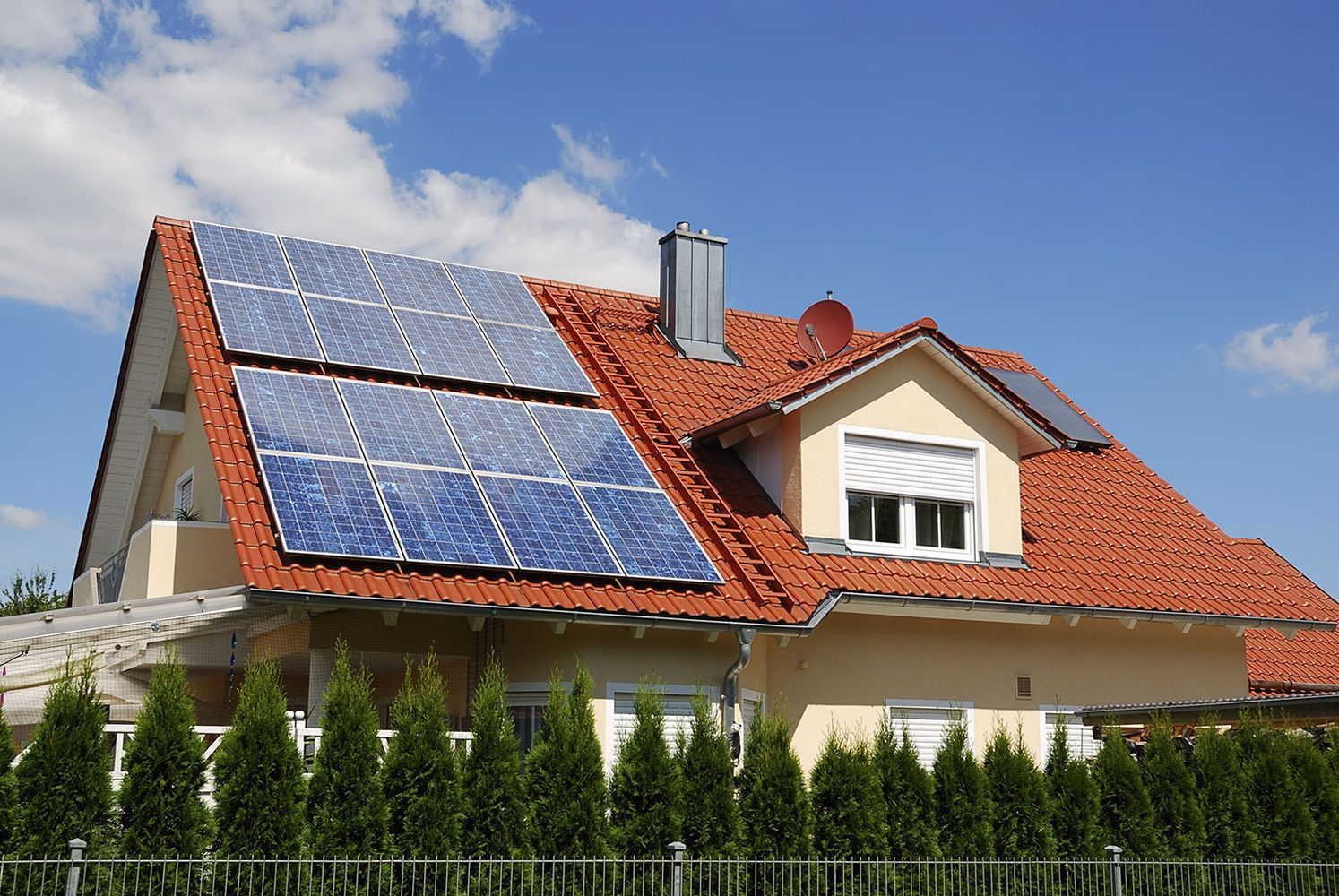 Haus mit Solarpaneelen auf dem Dach und roten Ziegeln hinter einer Hecke unter blauem Himmel