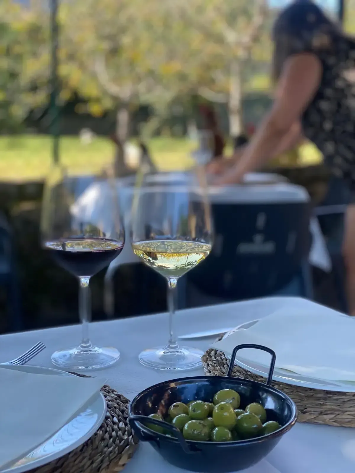 Dos copas de vino, una tinto y otra blanco, aceitunas y una mujer en una mesa al aire libre.