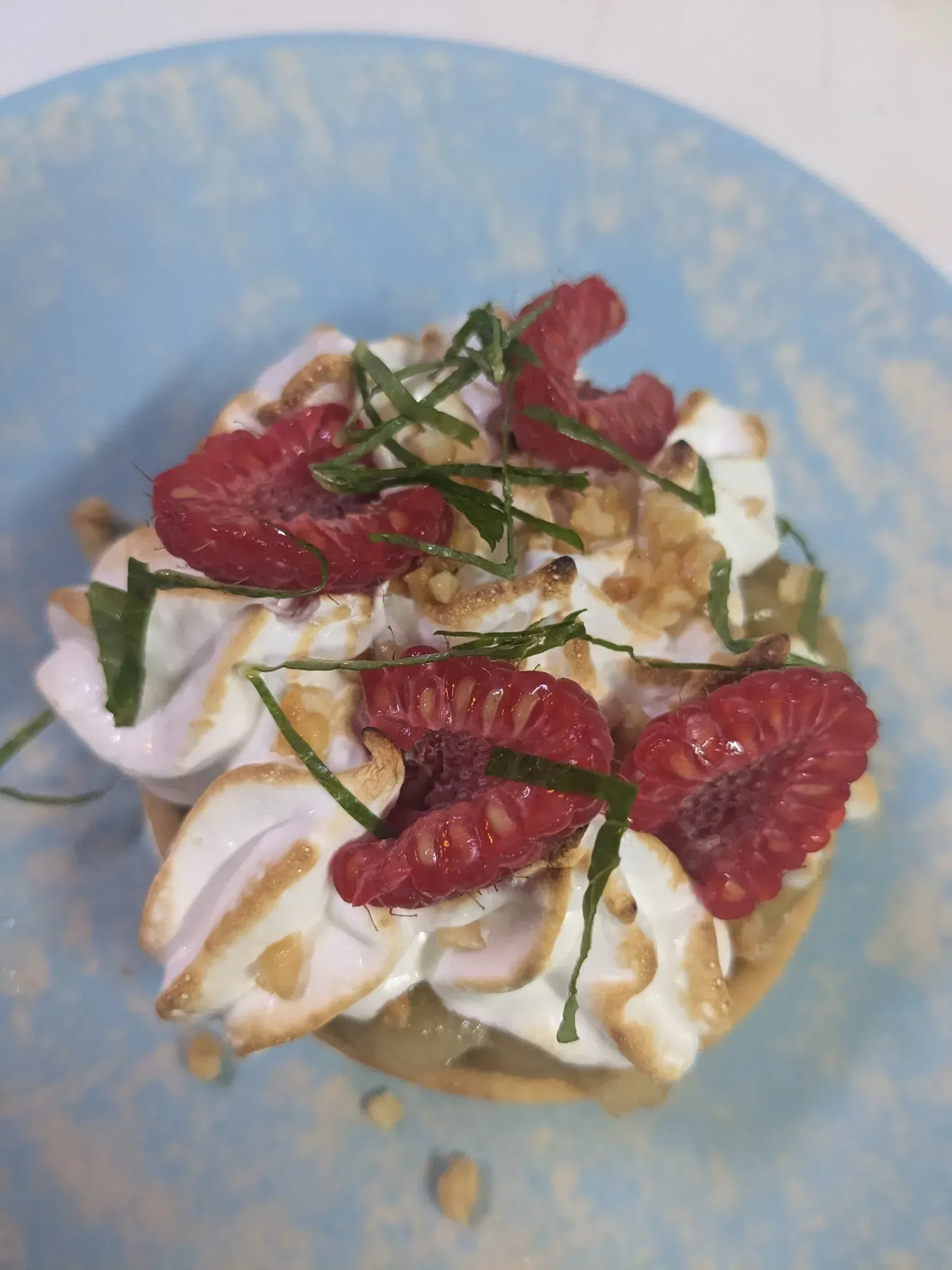 Tarta con merengue, frambuesas y guarnición de hierbas en plato azul.