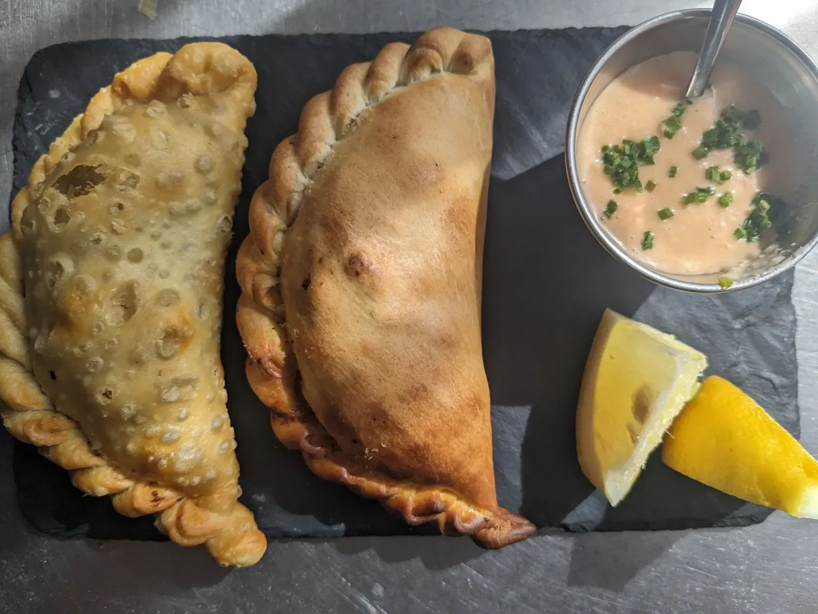 Dos empanadas doradas, salsa en un tazón pequeño y una rodaja de limón sobre una pizarra negra.