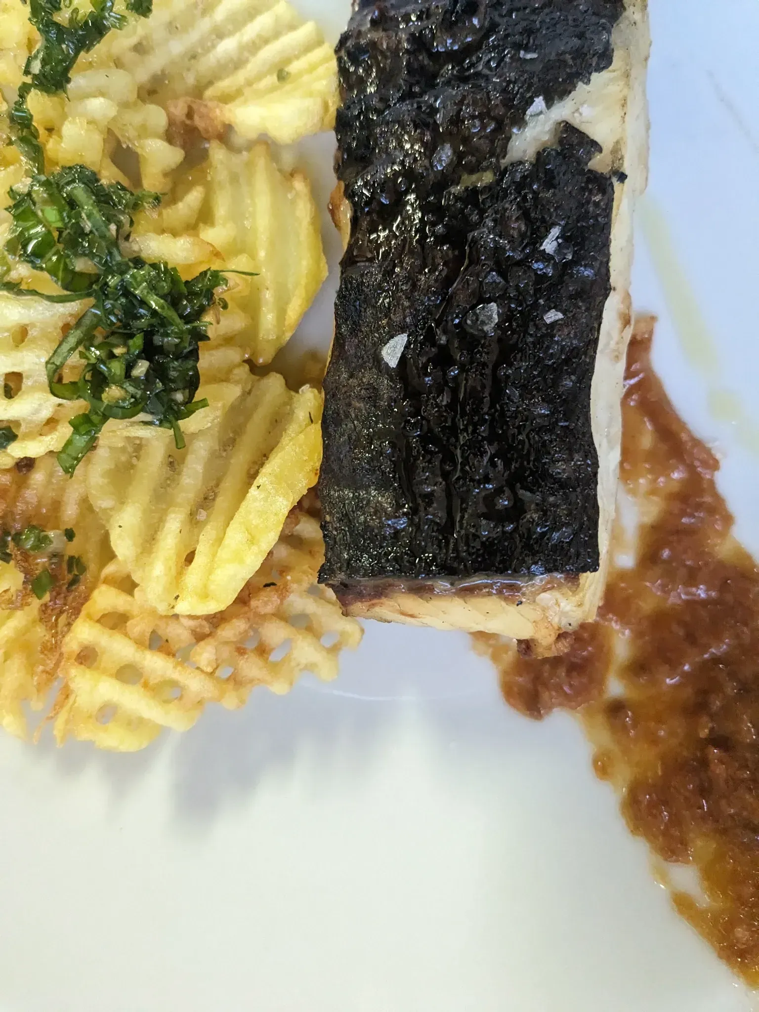 Plato con pescado cubierto con costra oscura, patatas onduladas y salsa.