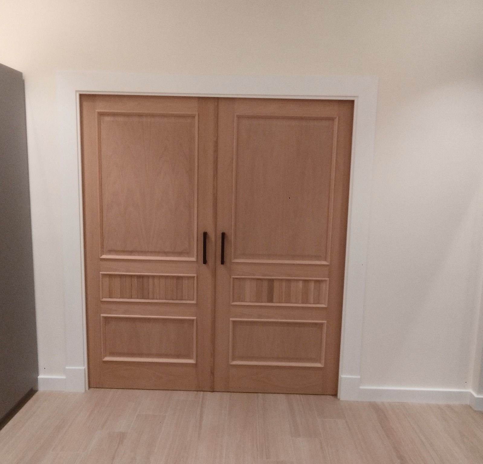 Puertas dobles de madera con tiradores negros enmarcadas en una pared blanca del pasillo.