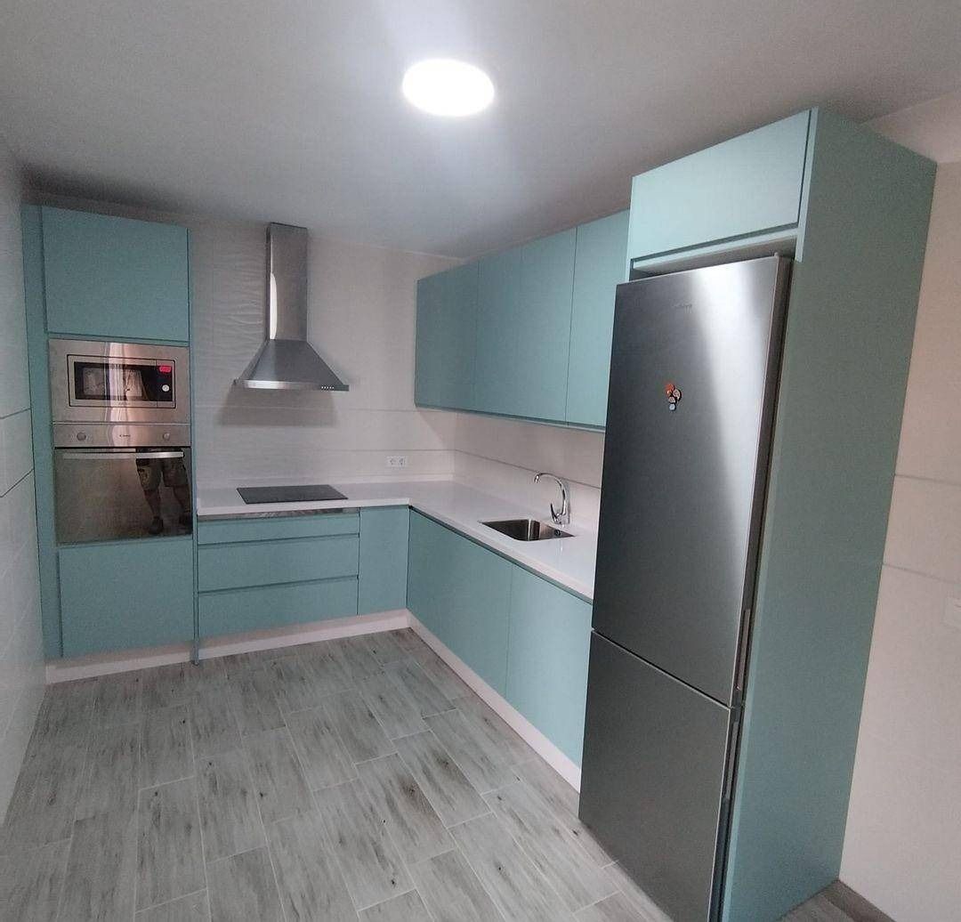 Cocina moderna en forma de L con armarios de color verde azulado, electrodomésticos de acero inoxidable y suelo gris claro.