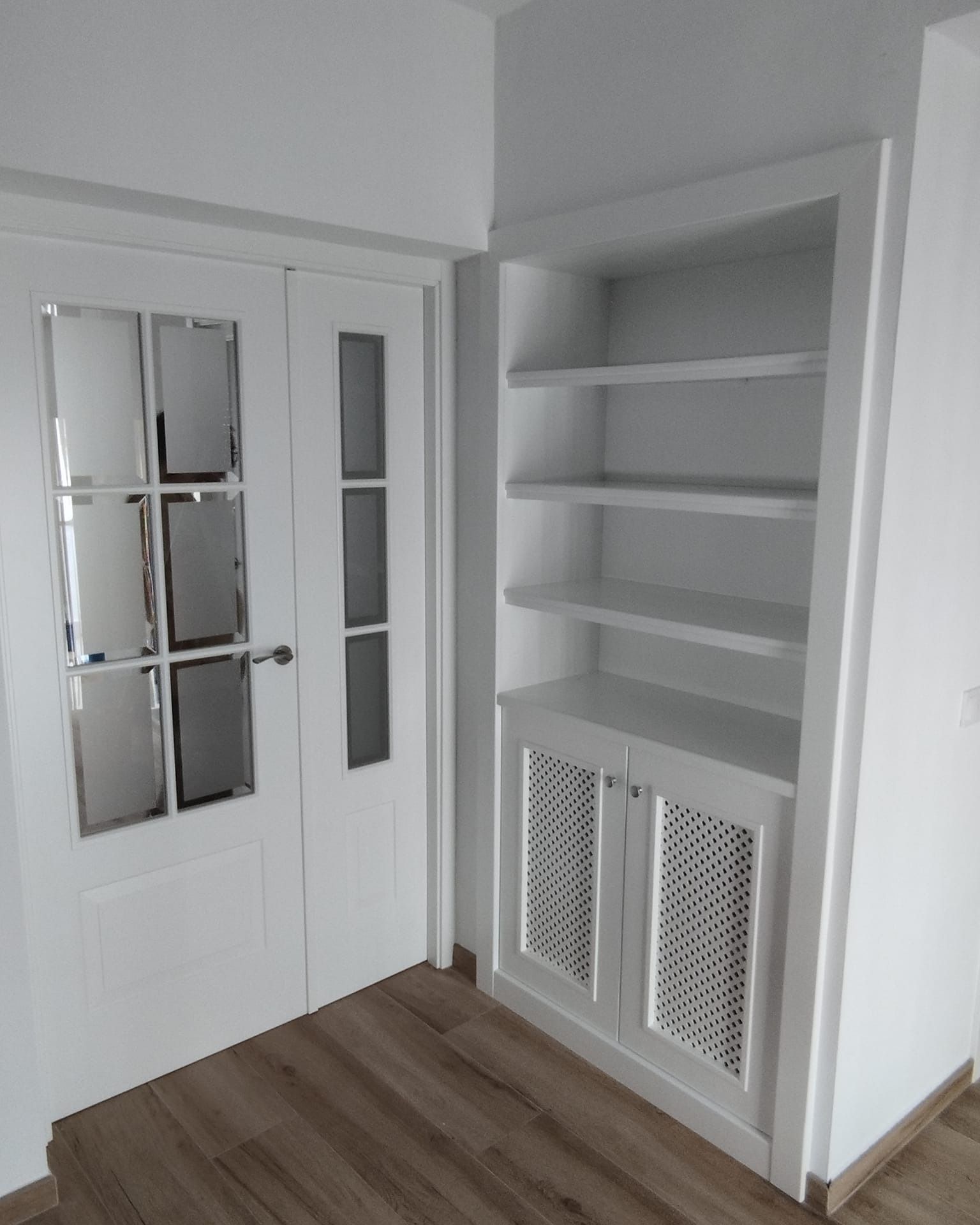 Estanterías y armario blancos empotrados junto a una puerta blanca en una habitación con suelo de madera.