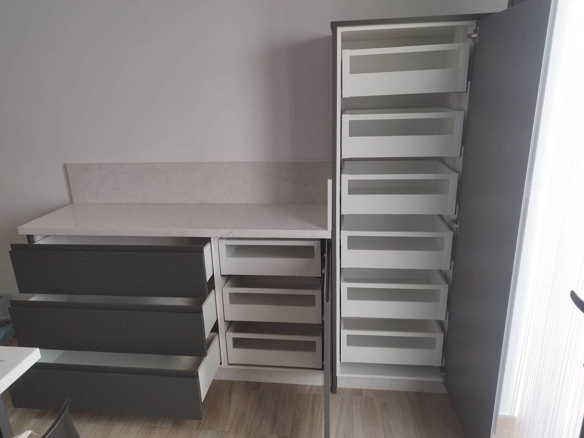 Pequeña oficina con escritorio oscuro, encimera clara y cajonera blanca alta contra una pared gris.