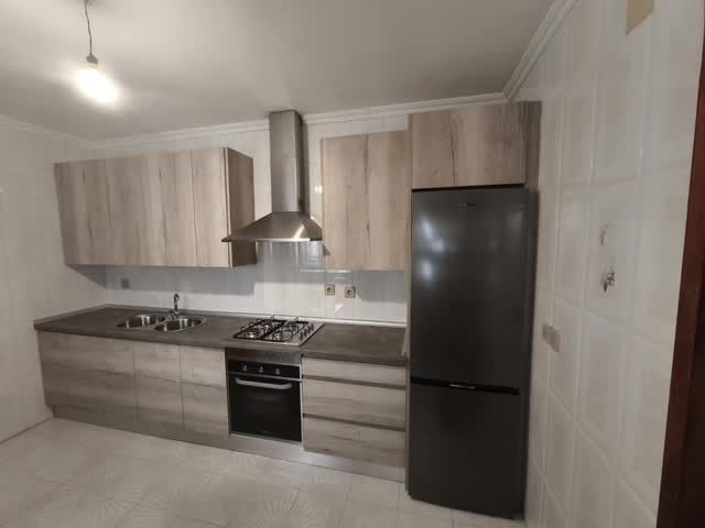 Cocina moderna con gabinetes de madera clara, encimeras grises, campana extractora de acero inoxidable y refrigerador negro.