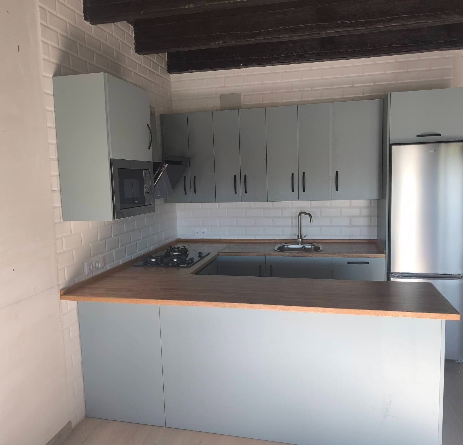 Pequeña cocina moderna con armarios grises, encimera de madera, fregadero, fogones y frigorífico de acero inoxidable.